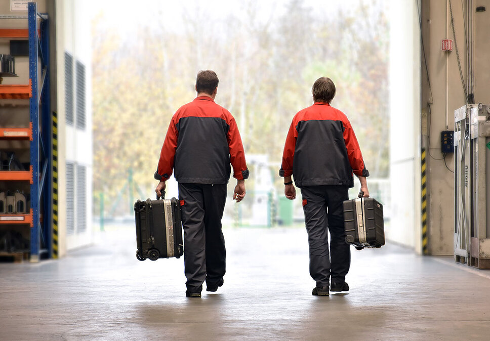 Two employees from weba's mobile toolmaking service, dressed in workwear and carrying toolboxes, are moving through a factory hall. They are on their way to install or maintain forming and stamping tools on-site at the customer's location.