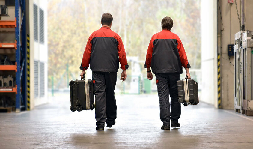 Zwei Mitarbeiter des mobilen Werkzeugbau-Services von weba in Arbeitskleidung, die mit Werkzeugkoffern in einer Werkshalle unterwegs sind. Sie sind auf dem Weg zur Installation oder Wartung von Umform- und Stanzwerkzeugen vor Ort beim Kunden.