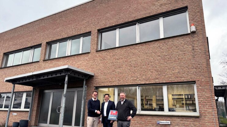 Strategic Expansion: weba Strengthens Innovation in Germany Three men standing in front of a brick building, two shaking hands while the third holds a document with weba branding.