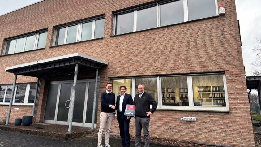 Drei Männer stehen vor einem Backsteingebäude, zwei schütteln sich die Hand, während der dritte ein Dokument mit weba-Branding hält.