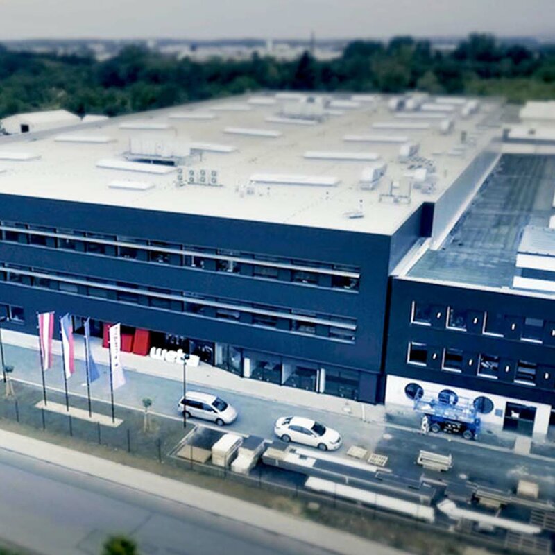 weba Olomouc - Serienproduzent und Experte in der Warmumformung Luftaufnahme des Produktionswerkes von weba für Warmumformung und Serienfertigung in Olomouc. Die Aufnahme zeigt die anthrazitfarbene Gebäudefront und dahinter eine riesige Produktionshalle. Vor dem Werk wehen die österreichische, tschechische, europäische sowie die Unternehmensflaggen.