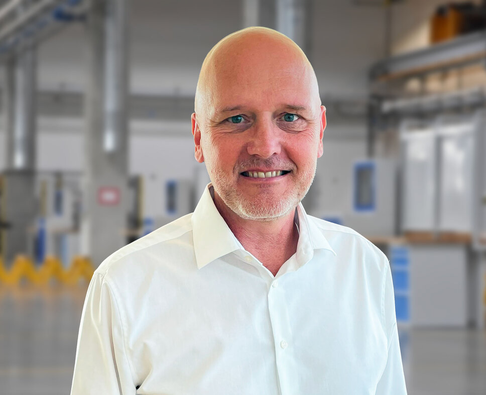 Portrait of Andreas Mehlmayer in a modern industrial setting. He is wearing a white shirt and smiling at the camera. The blurred background shows bright manufacturing equipment and machinery, reflecting a clean and precise production environment.