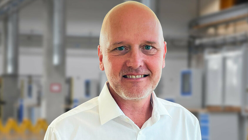 Porträt von Andreas Mehlmayer in einem modernen Industrieumfeld. Er trägt ein weißes Hemd und lächelt in die Kamera. Im unscharfen Hintergrund sind helle Fertigungseinrichtungen und Maschinen erkennbar – typisch für eine präzise und saubere Produktionsumgebung.