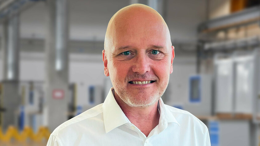 Porträt von Andreas Mehlmayer in einem modernen Industrieumfeld. Er trägt ein weißes Hemd und lächelt in die Kamera. Im unscharfen Hintergrund sind helle Fertigungseinrichtungen und Maschinen erkennbar – typisch für eine präzise und saubere Produktionsumgebung.