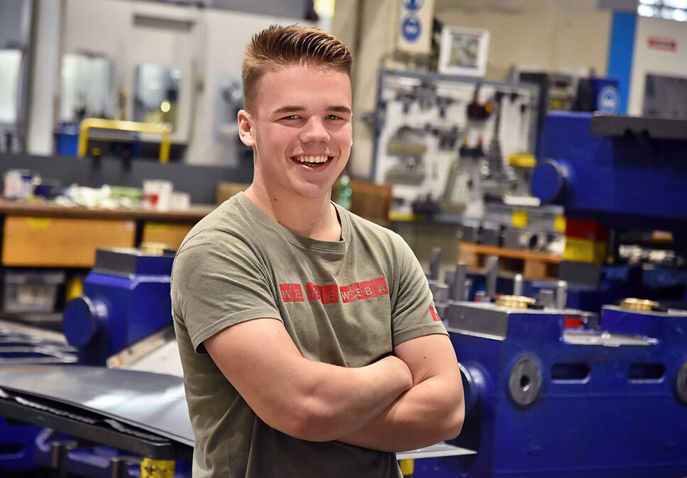 Jobs in Steyr - Lehre bei weba Werkzeugbau als Werkzeugbautechniker Ein Lehrling bei weba Werkzeugbau, fotografiert in der Produktionshalle, lächelt direkt in die Kamera. Er trägt ein T-Shirt mit der Aufschrift 'We are WEBA', was seinen Stolz und seine Zugehörigkeit zum Unternehmen zum Ausdruck bringt. Im Hintergrund sind mehrere Werkbänke und Teile eines dunkelblauen Umformwerkzeugs sichtbar, was auf die praktische und lernintensive Umgebung hinweist, in der der Lehrling ausgebildet wird. Das Bild strahlt Jugendlichkeit, Optimismus und das Engagement des Lehrlings für seine Ausbildung aus, während es gleichzeitig die unterstützende und lehrreiche Atmosphäre bei weba hervorhebt. Das Bild steht symbolisch für Lehrstellen Jobs in Steyr bei weba Werkzeugbau.