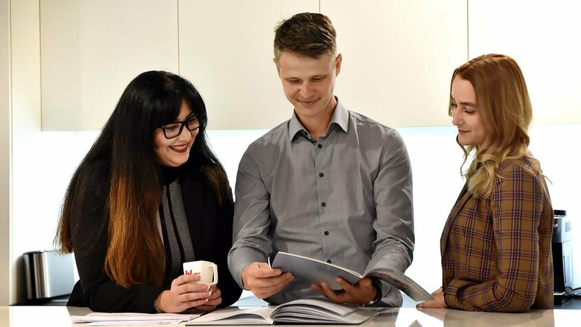 Drei junge Berufstätige im Büro tauschen sich über Unterlagen aus und diskutieren fachliche Inhalte – Beispiel für Hard Skills und Soft Skills.