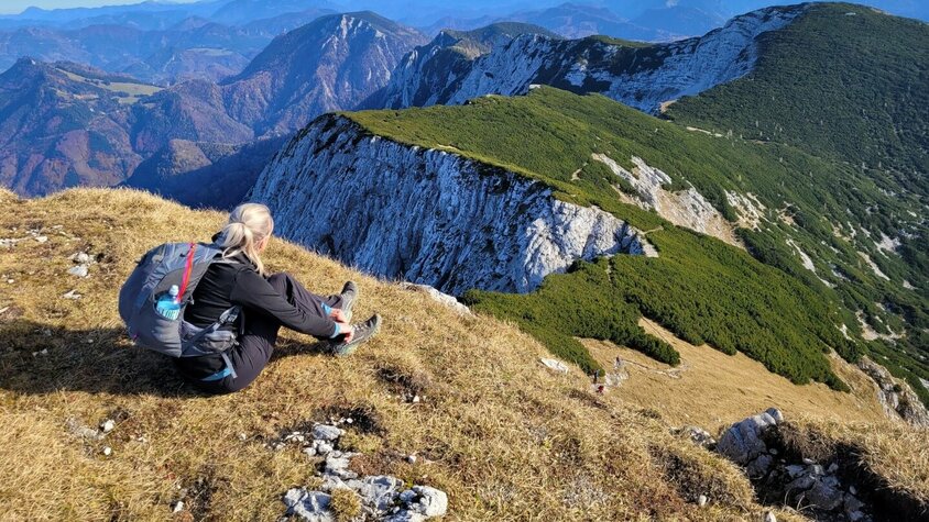 Bettina Gmainer mit Rucksack sitzt auf einer grasbewachsenen Anhöhe und blickt auf eine beeindruckende Berglandschaft mit grünen und felsigen Gipfeln.