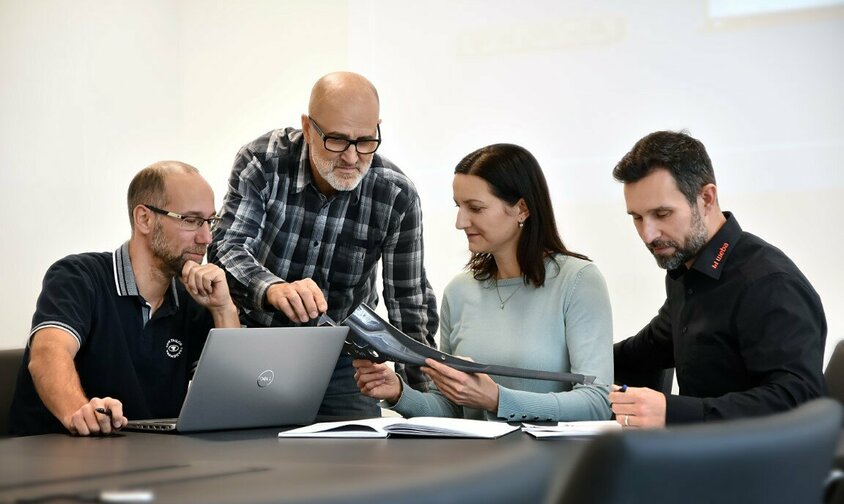 Corporate Social Responsibility - Corporate Policy  Three male employees and one female employee of weba Werkzeugbau are sitting in a meeting discussing a steel component. A laptop and documents are on the table.