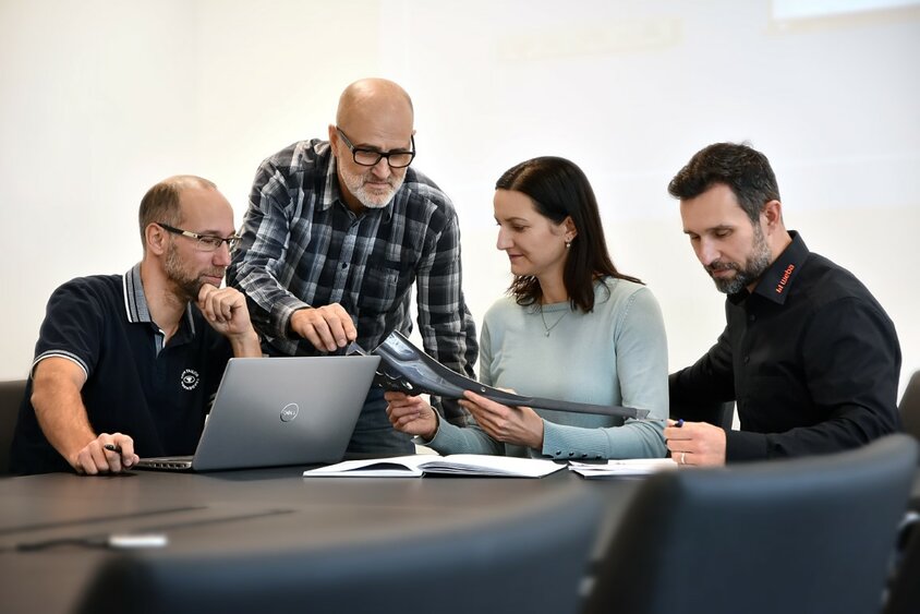 weba Werkzeugbau - Employee Blog Three male and one female employee of weba Werkzeugbau are sitting in a meeting discussing a steel component. A laptop and documents are on the table.