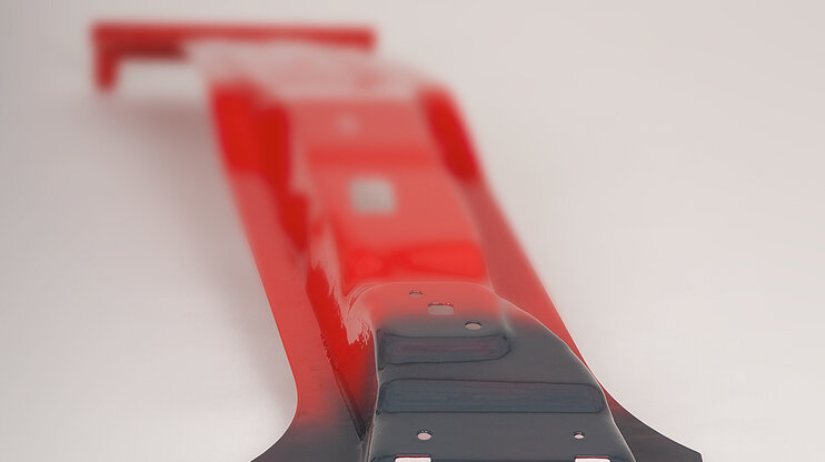 A B-pillar, manufactured from ultra-high-strength material, is shown in a striking red. A SoftZone, specifically developed for customized crash performance, is prominently highlighted in a deep anthracite grey, clearly standing out from the rest of the pillar. The color contrast emphasizes the unique properties of the B-pillar and the technical innovation behind the development of tailored safety features.