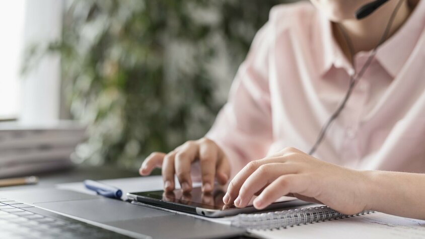 Das Bild zeigt eine Mitarbeiterin im Kundenservice, die mit einem Headset ausgestattet ist und parallel an einem Tablet sowie einem Laptop arbeitet. Sie macht sich Notizen in einem Notizbuch und zeigt volle Konzentration. Im Hintergrund sind grüne Pflanzen und Akten zu sehen, was auf eine moderne und organisierte Arbeitsumgebung hindeutet.