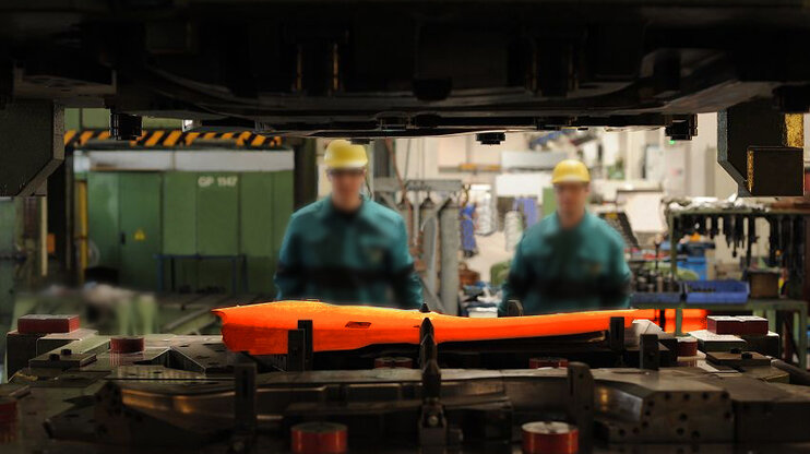 Glühende Stahlplatine in Werkzeug für das Presshärten mit Produktion im Hintergrund und verschwommenen Werkzeugbautechnikern