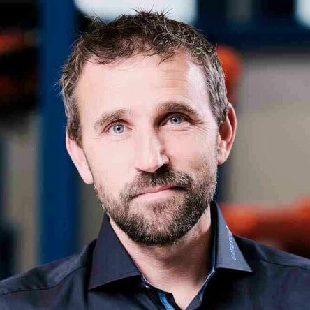 Herr Fiebiger, Betriebsleiter bei Gatzsch Schweißtechnik, steht in einem industriellen Umfeld neben einem technischen Gerät. Er trägt ein dunkelblaues Hemd und eine Jeans, eine Hand locker in der Hosentasche, während die andere Hand auf dem Gerät ruht. Im Hintergrund sind industrielle Anlagen und Maschinen zu sehen.