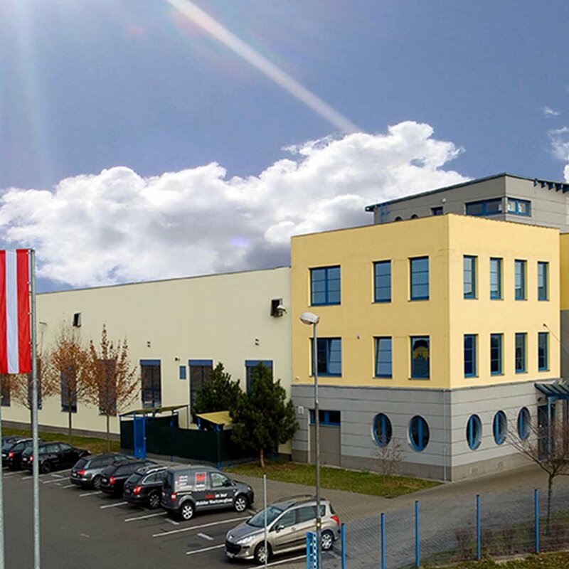 weba Olomouc - Experte in der Warmumformung Seitenaufnahme des Gebäudes des Produktionsstandorts Olomouc aus den Gründungszeiten. Strahlendblauer Himmel mit weißen Wolken. Das Gebäude ist in gelb-grauer Farbe gehalten, und davor wehen die Flaggen der Europäischen Union, Tschechiens (CZ) und Österreichs (AT).