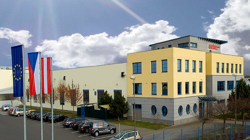 Seitenaufnahme des Gebäudes des Produktionsstandorts Olomouc aus den Gründungszeiten. Strahlendblauer Himmel mit weißen Wolken. Das Gebäude ist in gelb-grauer Farbe gehalten, und davor wehen die Flaggen der Europäischen Union, Tschechiens (CZ) und Österreichs (AT).