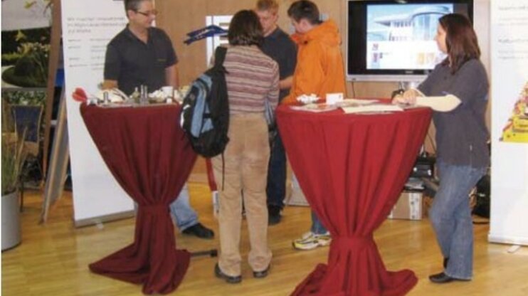 Personen stehen an einem Messestand von weba Werkzeugbau bei der Lehrlingsmesse 2008 in Steyr. Zwei Stehtische mit roten Hussen, Infomaterialien und Werkstücke sind ausgestellt, Besucher informieren sich bei den Mitarbeitern.