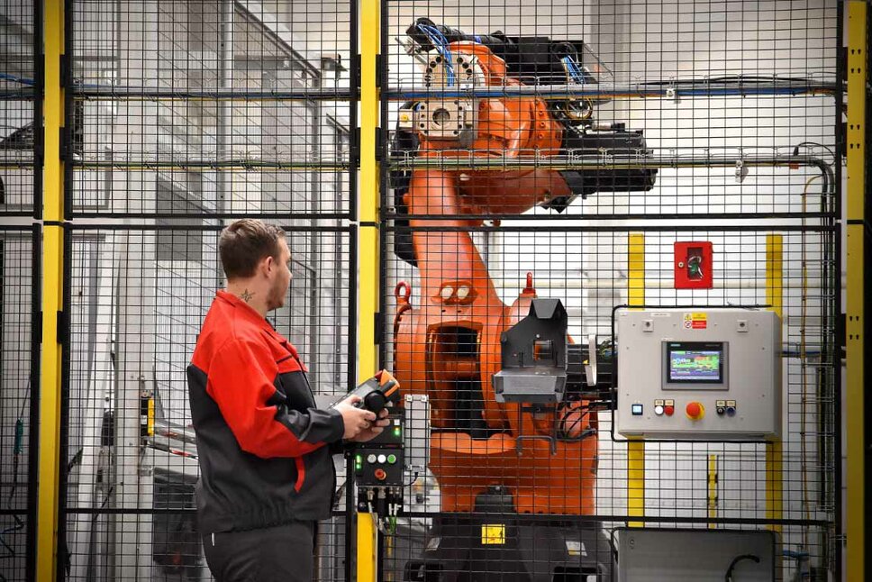 Operation of an Industrial Robot in Mechanical Machining An employee in red workwear is operating an industrial robot in a secured production cell. The robot arm is visible behind a safety fence, and the operator is holding a control device to monitor or adjust the robot's movements. Next to the robot, there is a control panel with a digital display used for managing and monitoring the robotic processes. The scene illustrates the use of automation and advanced robotics in mechanical machining.