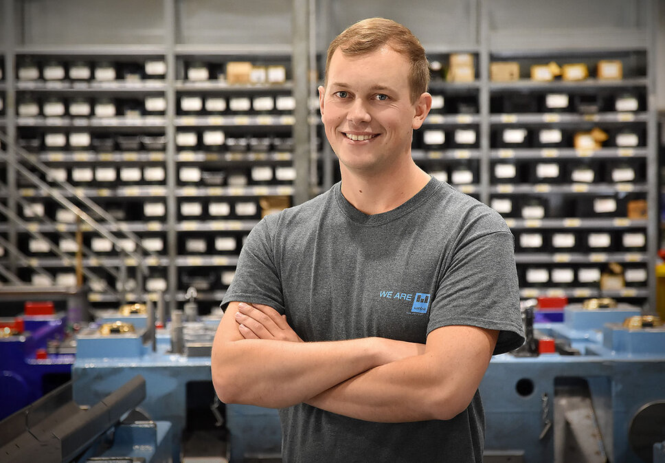 Jobs in Steyr - Praktikum bei weba Werkzeugbau Ein Werkzeugbautechniker von weba Werkzeugbau, fotografiert in der Produktionsumgebung, blickt direkt und mit einem stolzen Lächeln in die Kamera. Er steht in einer selbstbewussten Pose, die seinen Stolz auf seine Arbeit unterstreicht. Im Hintergrund sind ordentlich angeordnete Betriebsmittelregale und ein markantes blaues Umformwerkzeug zu sehen, was auf die gut organisierte und technologisch fortschrittliche Arbeitsumgebung hinweist. Das Bild vermittelt ein Gefühl von Professionalität und Zufriedenheit im Arbeitsumfeld, und hebt die persönliche Verbindung des Technikers zu seiner Arbeit und seinem Arbeitsplatz hervor. Das Bild steht symbolisch für Praktikum-Jobs in Steyr.
