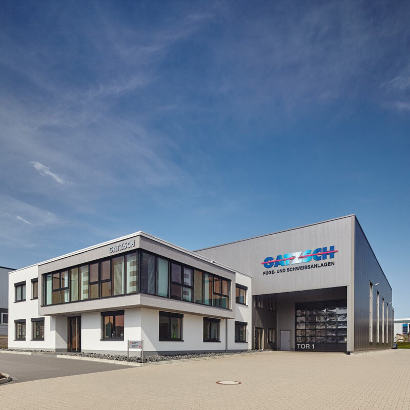 Gatzsch Schweißtechnik - Experte für High-Tech Fügeanlagen Die Außenansicht des Werkes Gatzsch Schweißtechnik GmbH zeigt ein strahlendblaues Himmel und das Verwaltungsgebäude im Fokus. Dahinter erstreckt sich die Produktionsanlage, auf der ein großes Logo mit den Worten 'Gatzsch Füge- und Schweissanlagen' deutlich sichtbar ist.