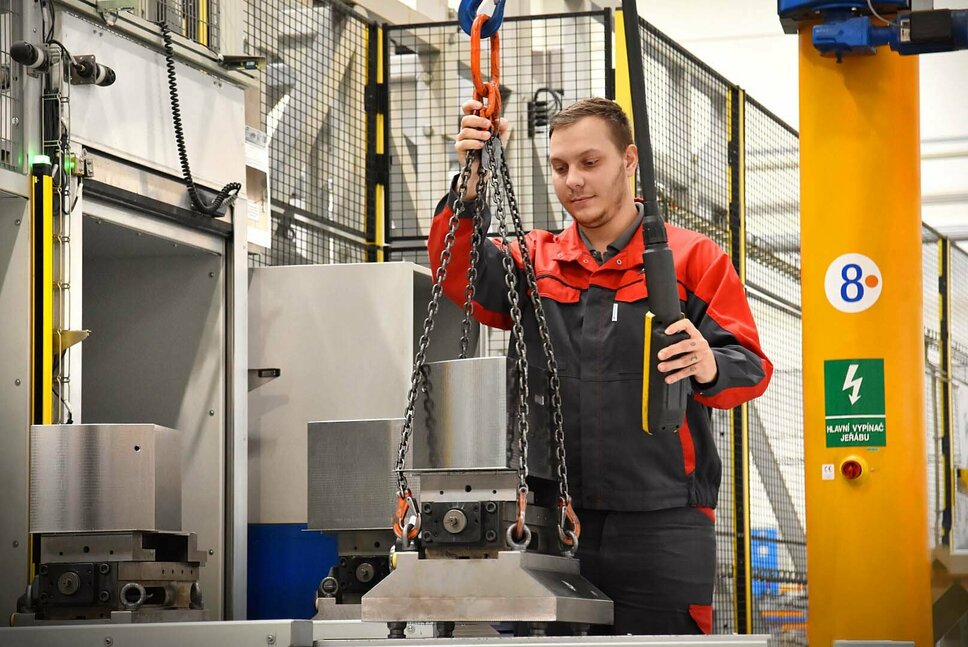 Přesné předběžné vybavení ve smluvní výrobě Obsluha stroje pracuje na přednastavovací stanici automatizovaného obráběcího centra pro smluvní výrobu. Pečlivě vkládá nástroje a součásti do seřizovacích stanic. Důraz je kladen na přesné umístění přesných nástrojů do určených pozic na složitém loži stroje. V pozadí je vidět průmyslové prostředí s vyspělou technologií.