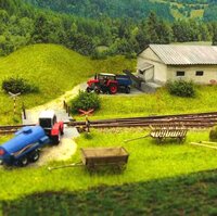 Detailně zpracovaná krajina modelové železnice s kolejemi, železničním přejezdem, traktory, přívěsy a zemědělskou budovou.