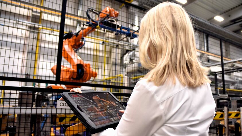 A woman stands in an industrial environment at weba Werkzeugbau in front of a large robotic gripper of a hot forming line. She holds a tablet in her hands, monitoring data or inputting commands for the robot. The focus is on the interaction between modern technology in tool manufacturing and parts production and human intervention.