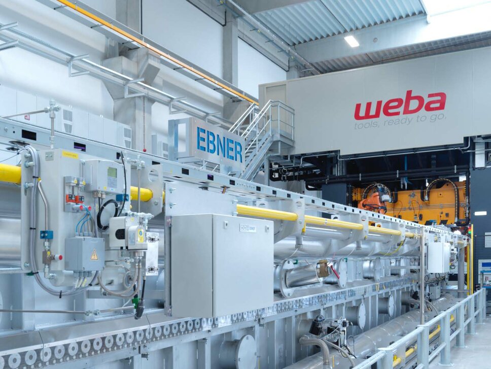 Photo of a hot forming system at weba Werkzeugbau, featuring a 22-meter-long roller hearth furnace and a 1,600-ton forming press. The system demonstrates industrial hot forming technology in action, where the furnace is used to heat metal sheets prior to forming, and the press shapes the heated metal into the desired form.