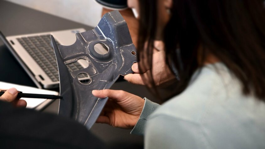 Foto einer Besprechung über die Qualitätskontrolle eines warmumgeformten Bauteils. Eine Frau hält das Bauteil in ihrer Hand, während eine andere Person auf eine bestimmte Stelle des Bauteils zeigt, um auf ein Detail hinzuweisen. Im Hintergrund des Bildes ist ein Teil eines Laptops und verschiedene Unterlagen sichtbar, was auf eine technisch detaillierte Analyse und Dokumentation des Prüfprozesses hinweist. Die Szene vermittelt einen Eindruck von Teamarbeit und dem Fokus auf Qualität in der Produktionsumgebung und der Lohnfertigung bei weba.
