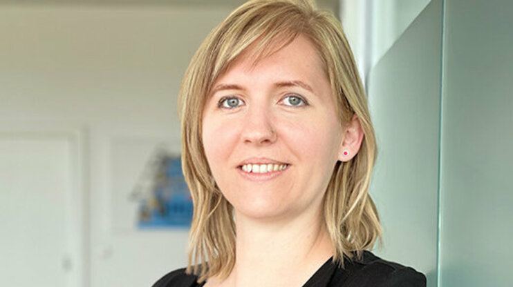 Portrait of Tanja Burghuber, HR Manager at weba Werkzeugbau, leaning casually against a frosted glass wall in Dietach, with a professional and friendly expression, highlighted by soft lighting and a blurred background. The photo is used in the contact section of the job openings for the Steyr location of weba Werkzeugbau.