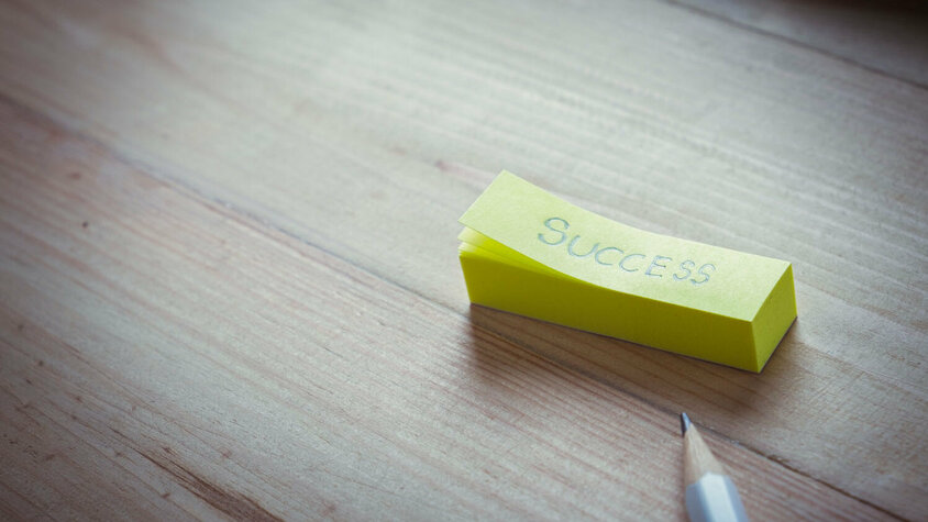 Gelber Notizzettel mit dem Wort „Success“ auf Holztisch, daneben ein Bleistift.