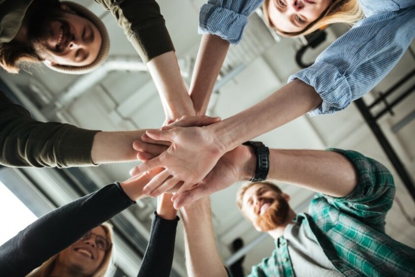 weba.inside - World of Work A young team smiles and stacks their hands in a circle. The image symbolizes weba’s workplace culture.