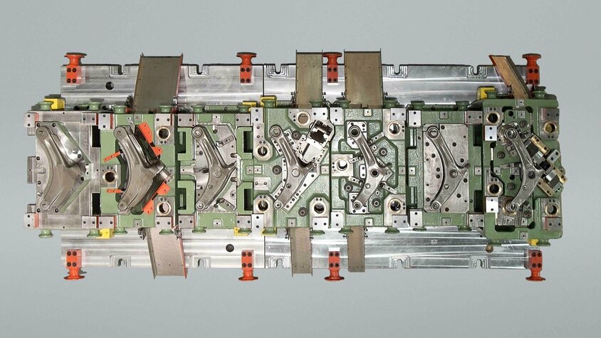 Top-Ansicht eines Transferwerkzeugs zur Herstellung eines Querlenkerarms; erkennbar sind mehrere Stationen zur Umformung und Positionierung des Bauteils in einem mehrstufigen Werkzeugaufbau.