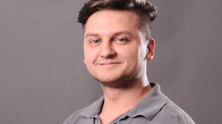 Portrét mladého muže s krátkými, na stranu oholenými vlasy a přátelským úsměvem. Na sobě má šedou polokošili a dívá se přímo do kamery. Pozadí je neutrální a šedé.