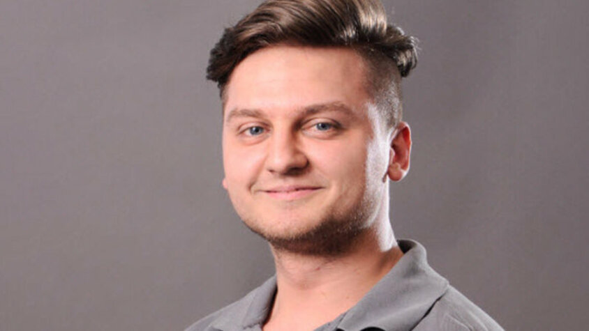 Porträt eines jungen Mannes mit kurzem, seitlich rasiertem Haar und einem freundlichen Lächeln. Er trägt ein graues Polohemd und blickt direkt in die Kamera. Der Hintergrund ist neutral und grau.