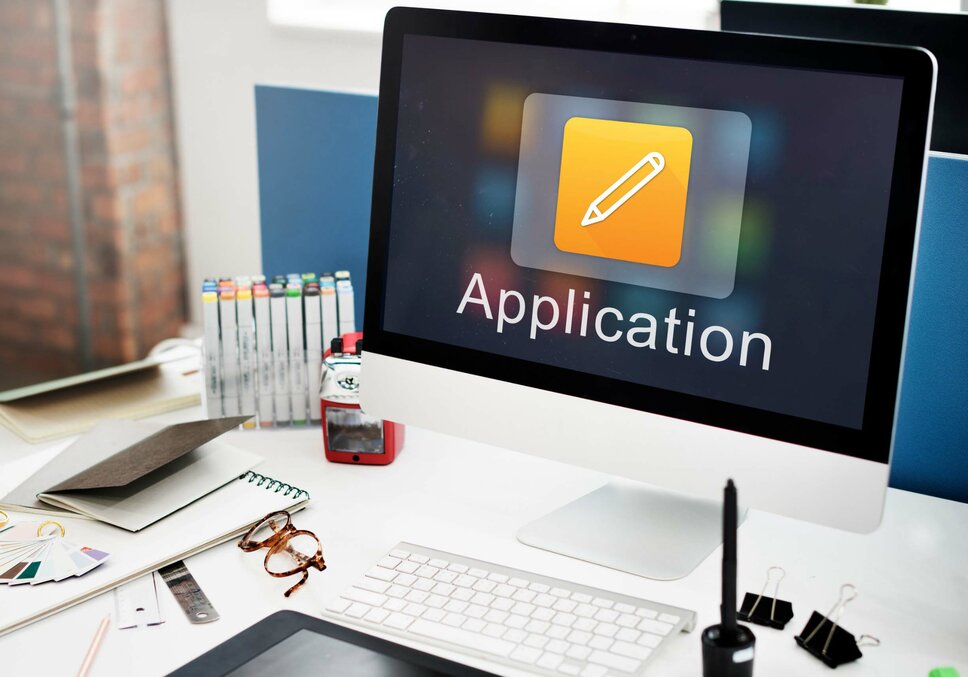 A desk with an Apple monitor, keyboard, and office supplies. The screen displays the word "Application," symbolizing job opportunities in Steyr at weba Werkzeugbau.