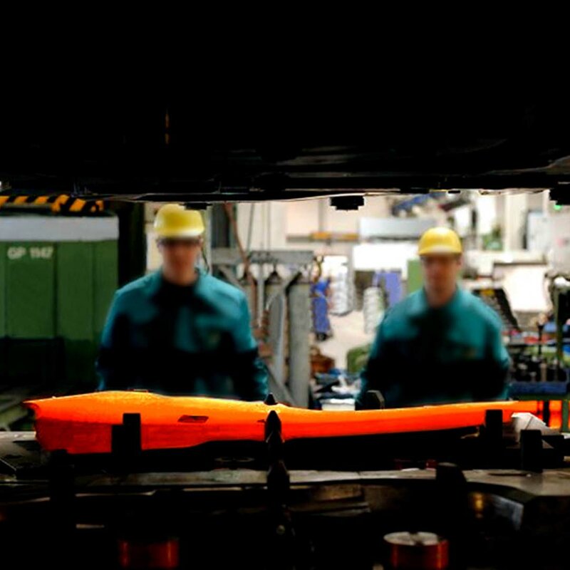 weba Werkzeugbau - Experte in der Warmumformung seit 1994 Ein durch den Spalt des Ober- und Unterteils einer Umformpresse aufgenommenes Bild fokussiert eine glühende Formplatine, die in ein Warmumformwerkzeug eingelegt ist. Im Hintergrund sind zwei Werkzeugbautechniker und die verschwommene Produktion zu sehen.