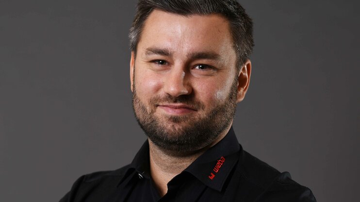 Alexander se prezentuje přátelsky a profesionálně: černá košile s červeným logem weba na šedém pozadí, usmívá se přímo do kamery.