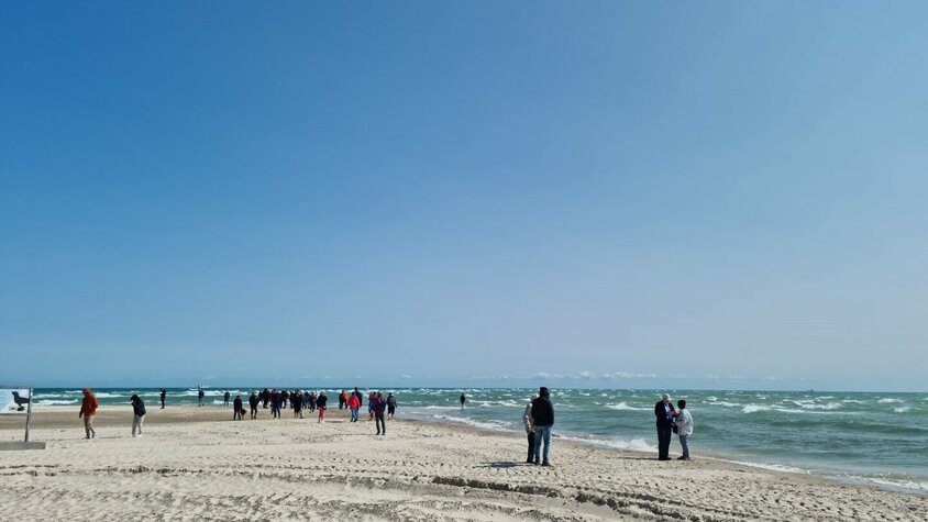 Eine Gruppe von Menschen spaziert bei sonnigem Wetter an einem Sandstrand entlang, wo sich Nord- und Ostsee treffen. Der Himmel ist strahlend blau, das Meer leicht bewegt.