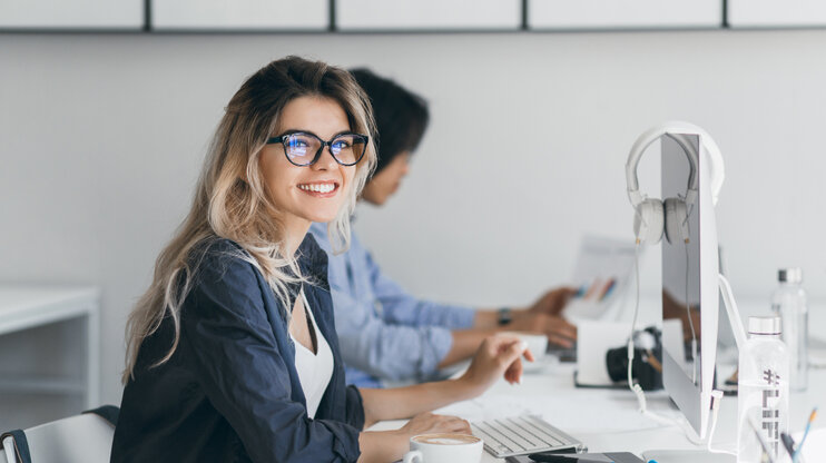 Young woman with glasses smiling at her desk in a modern office, working on a computer, with colleagues in the background.