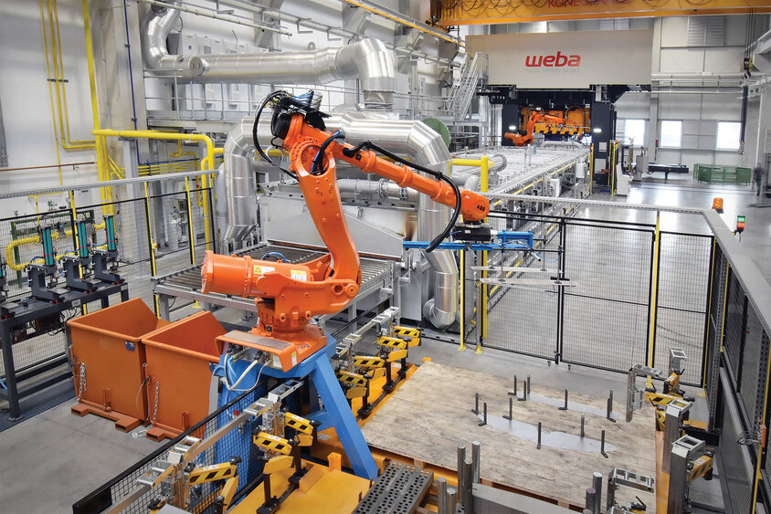 Foto einer Warmumformlinie, bestehend aus Transfergreifern, einem Durchlaufofen und einer Umformpresse. Deutlich sichtbar auf der Umformpresse ist das große Logo von weba.
