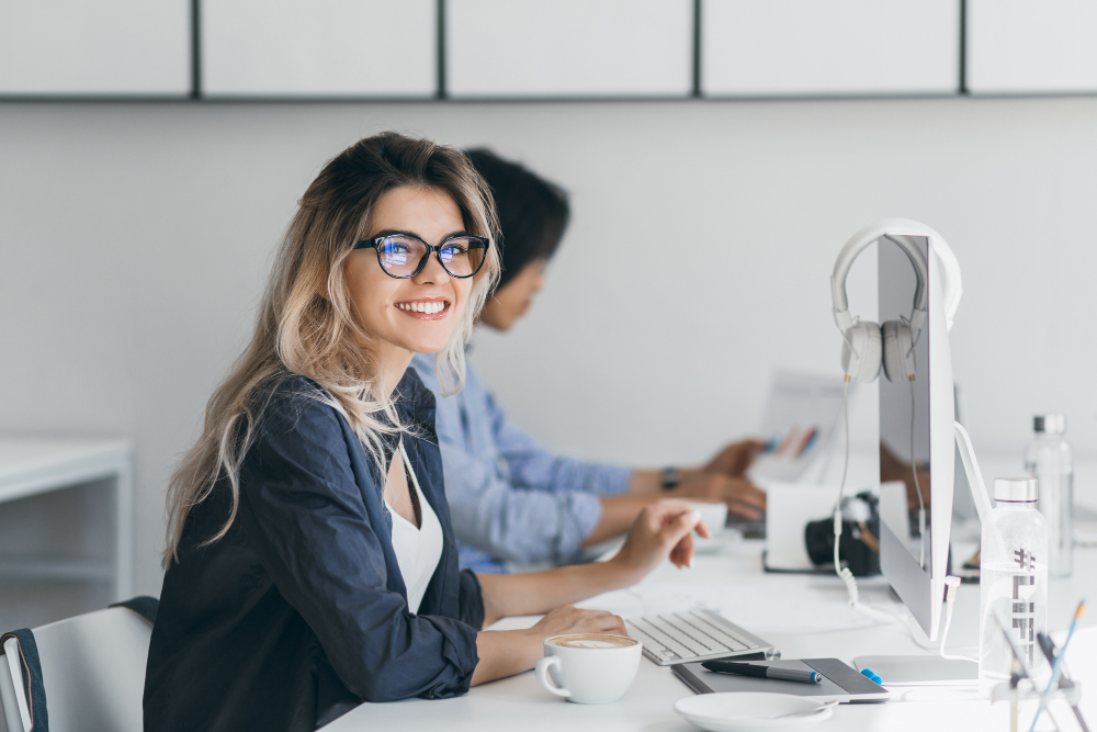 Young woman with glasses smiling at her desk in a modern office, working on a computer, with colleagues in the background.