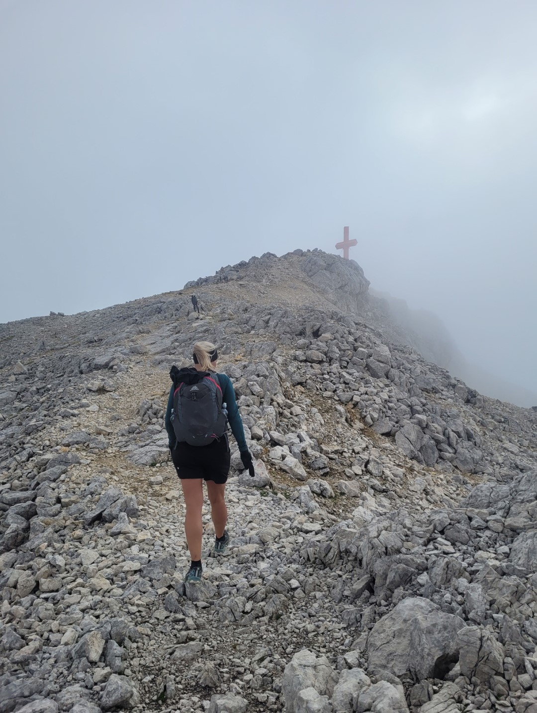 Bettina with a backpack climbs a rocky mountain path, approaching a summit cross. The sky is foggy, creating a rugged and adventurous atmosphere.