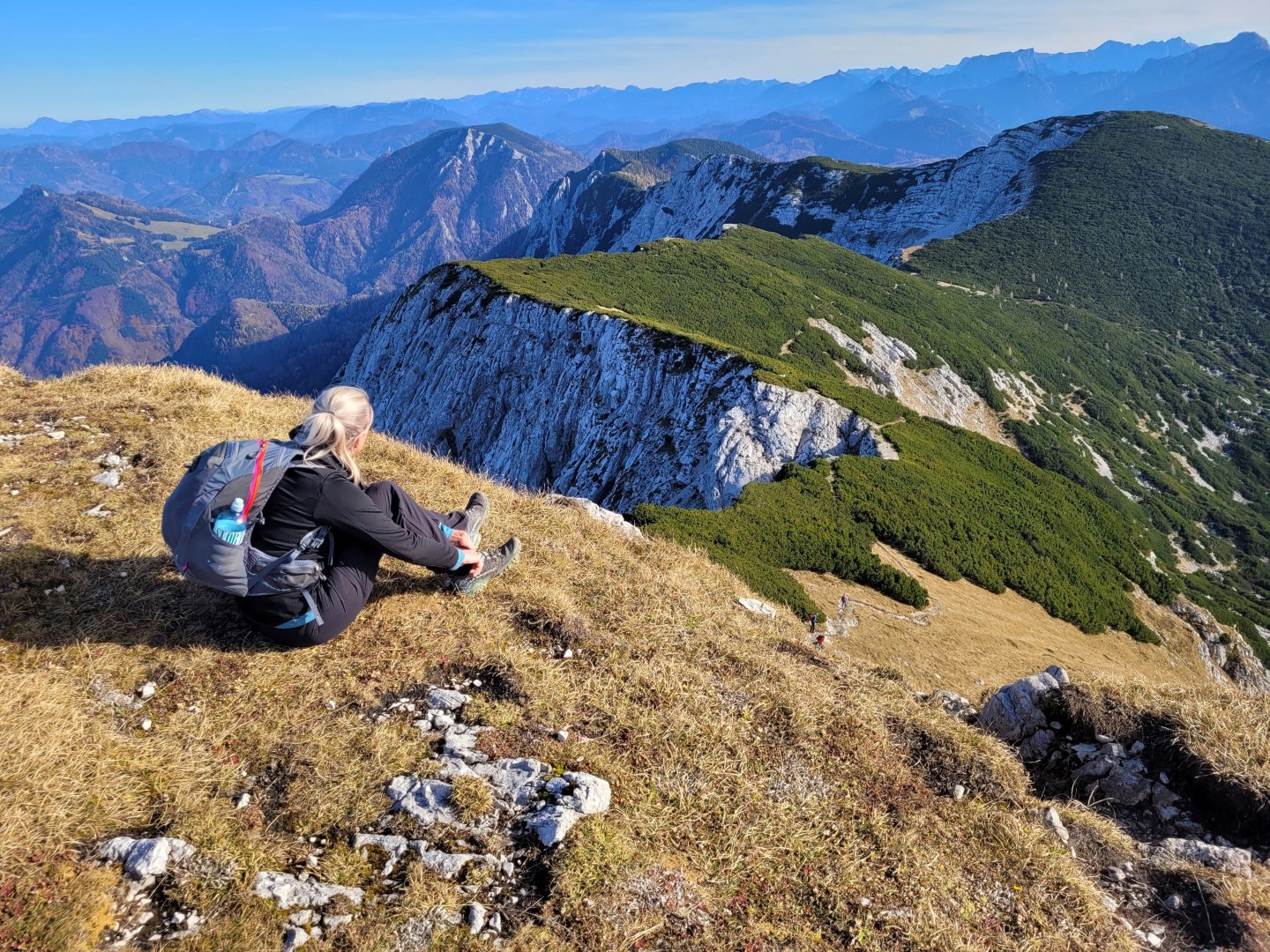 Bettina Gmainer with a backpack sits on a grassy hill, gazing at a breathtaking mountain landscape with green and rocky peaks.