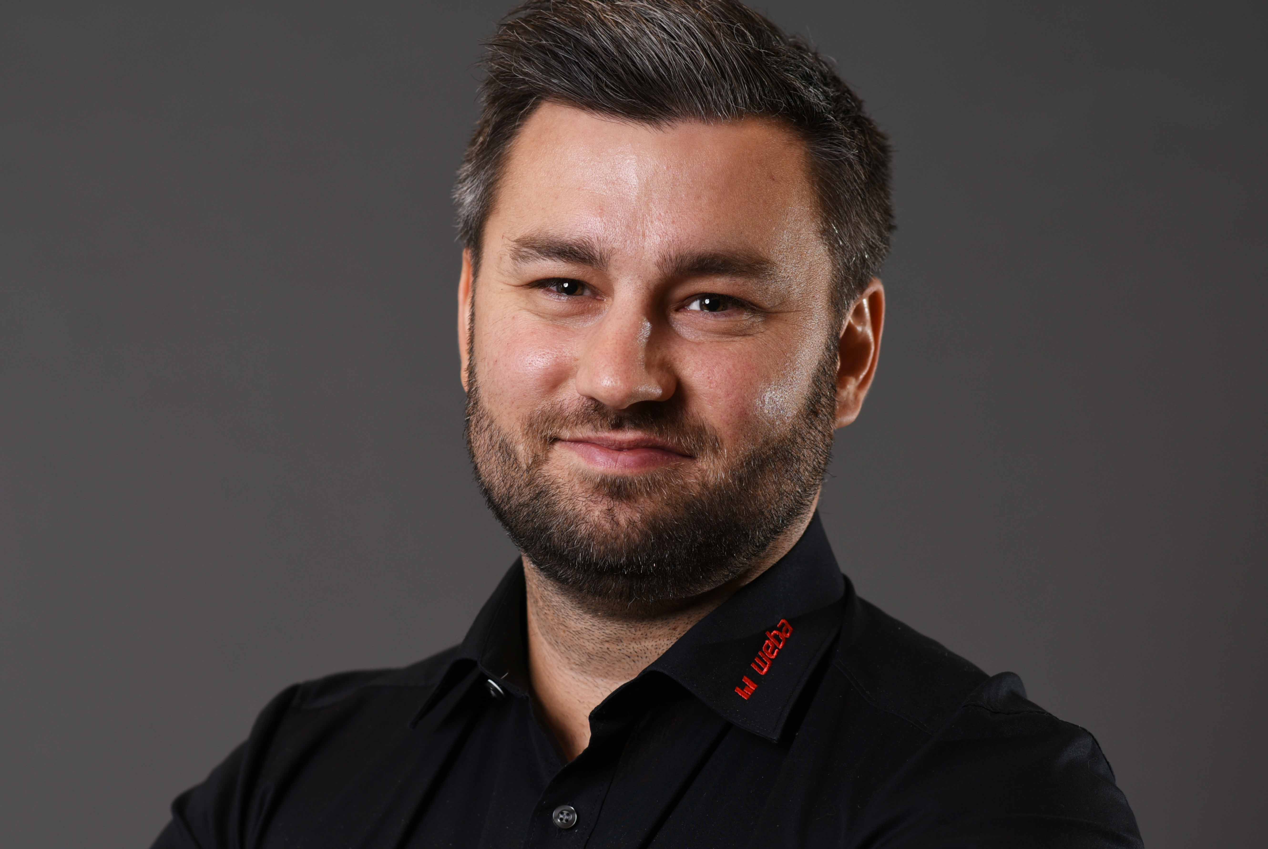 Alexander smiles confidently at the camera, wearing a black shirt with a red weba logo on the collar, posing in front of a neutral grey background.